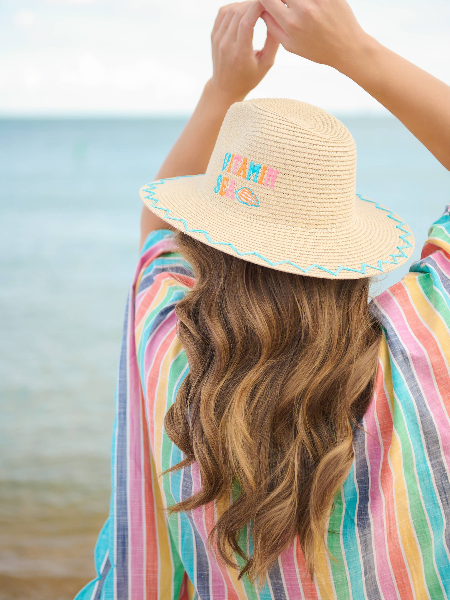 Shiraleah Vitamin Sea Sun Hat shown from back, worn by model at beach, straw hat with colorful embroidery and teal zigzag brim trim.