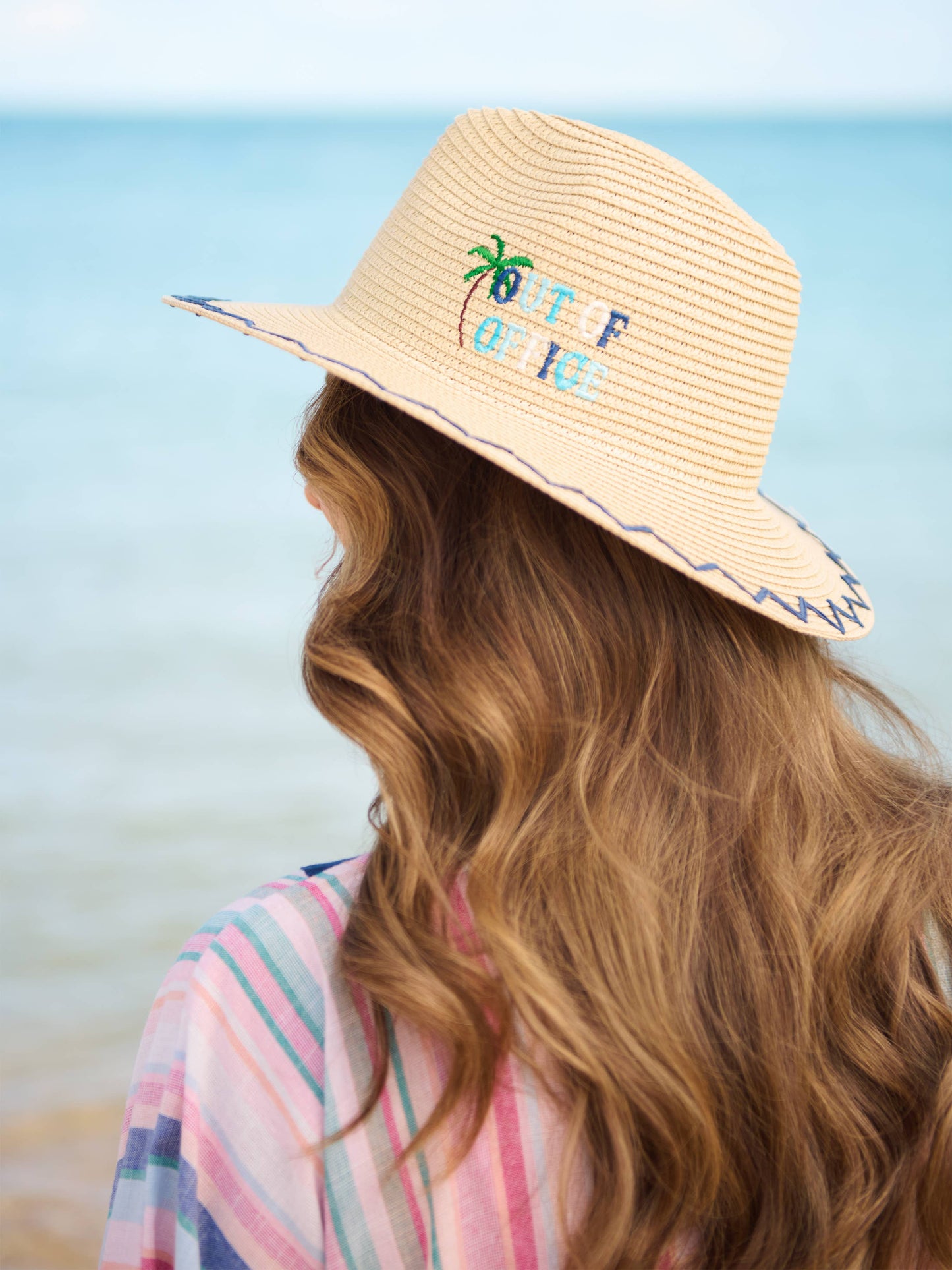 Out Of Office straw sun hat with embroidered palm tree and blue "Out Of Office" text, worn at the beach.