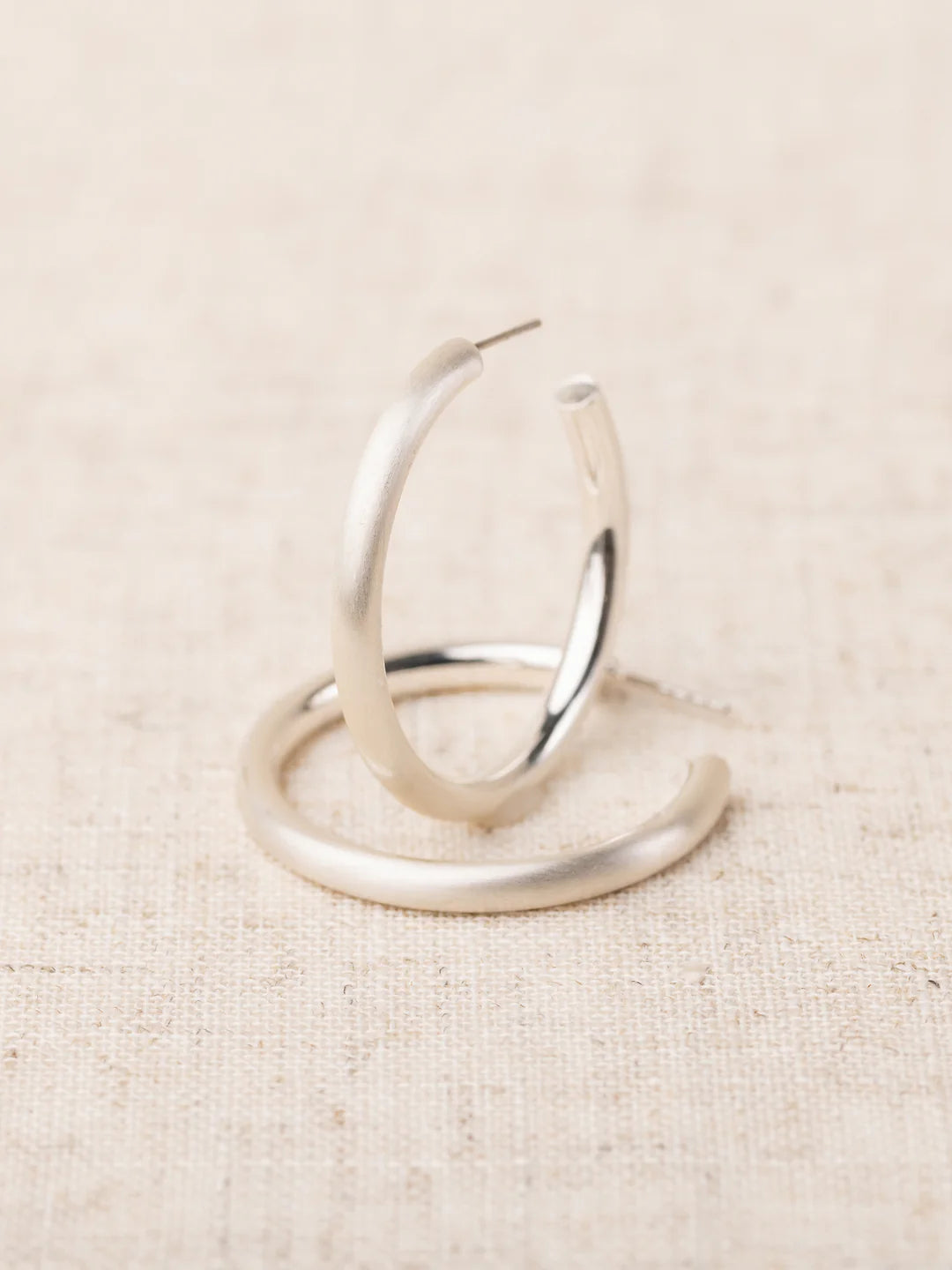 Heather silver hoop earrings with matte finish, one standing upright and one lying flat on linen fabric.