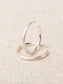 Heather silver hoop earrings with matte finish, one standing upright and one lying flat on linen fabric.